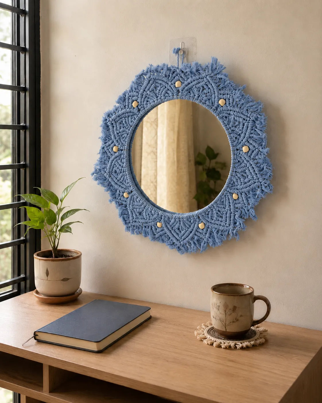 Round blue macrame mirror on a wall above a wooden desk with a plant, book, and mug.